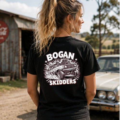 A woman wearing a black graphic t-shirt with a white and pink 'BOGAN SKIDDERS' print on the back, standing outdoors in front of a rustic barn and a vintage car.